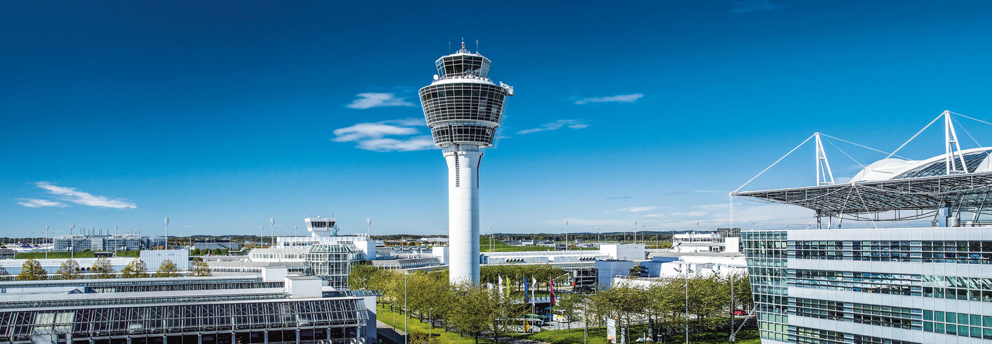 Integrierter Bericht 2017 Der Tower des Flughafens München erhebt sich vor strahlend blauem Himmel, flankiert von den markanten Terminaldächern. Die klare Darstellung steht für moderne Flughafeninfrastruktur und internationales Reisen.