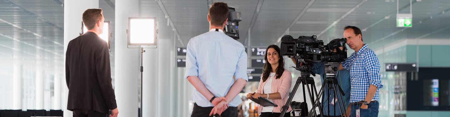 Medienproduktion am Flughafen München Kamerateam mit Beleuchtung und zwei Filmkameras in einem Terminal des Flughafens München; eine Person steht für eine Aufnahme vor der Kamera.