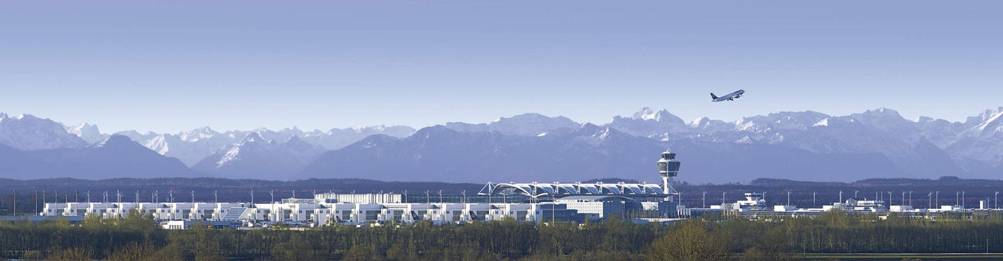 Flughafen München vor Alpenkulisse Panorama des Flughafens München mit Tower und Terminals vor den Alpen; ein Flugzeug steigt im Hintergrund auf.