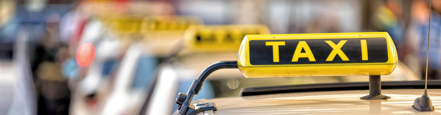 Taxi-Service am Flughafen München Nahaufnahme des gelben Taxi-Dachschilds mit schwarzer Aufschrift „TAXI“ auf einem Fahrzeug am Flughafen München; im Hintergrund sind unscharf weitere Fahrzeuge und Menschen zu erkennen, die Atmosphäre wirkt geschäftig, aber geordnet.