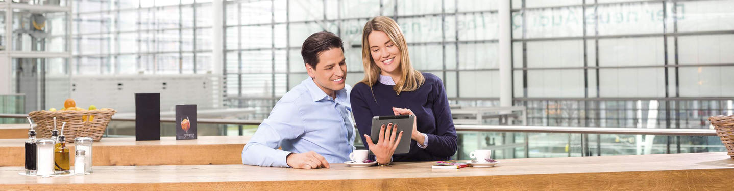 Service & Auskunft am Flughafen München Zwei Personen sitzen in einem modernen Flughafenbereich an einem Tresen und betrachten gemeinsam ein Tablet, neben ihnen stehen Tassen und kleine Gegenstände; die Szene steht sinnbildlich für Service und Auskunft am Flughafen München und wirkt freundlich, hell und einladend.