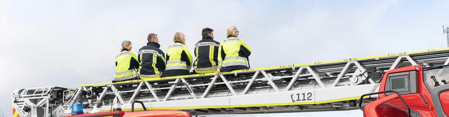 Ausbildung Werkfeuerwehr am Flughafen München Mehrere Auszubildende der Werkfeuerwehr in Warnjacken sitzen auf einer ausgefahrenen Drehleiter eines Feuerwehrfahrzeugs am Flughafen München; ruhige, beobachtende und einsatzbereite Atmosphäre.