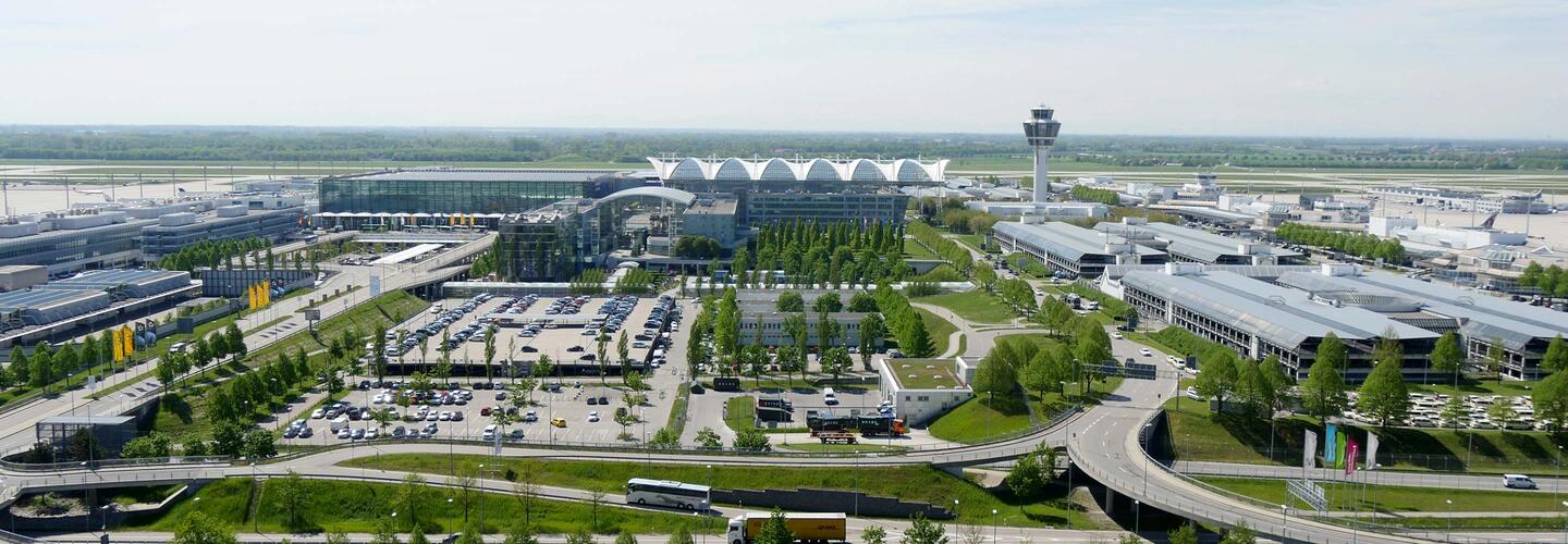 Panoramic view at Munich Airport Wide panoramic view of Munich Airport with green open spaces, modern terminal buildings, and the control tower in the background. The scene appears clear, expansive, and structured—a lively, international transportation hub in sunny weather.