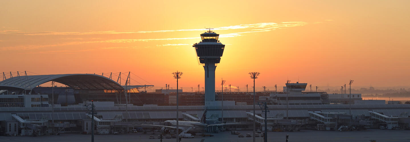Panoramic view on Munich Airport The tower of Munich Airport stands out against an intense orange evening sky. Silhouettes of terminal roofs and lighting masts stand out against the warm light. The atmosphere is calm, atmospheric, and slightly melancholic.