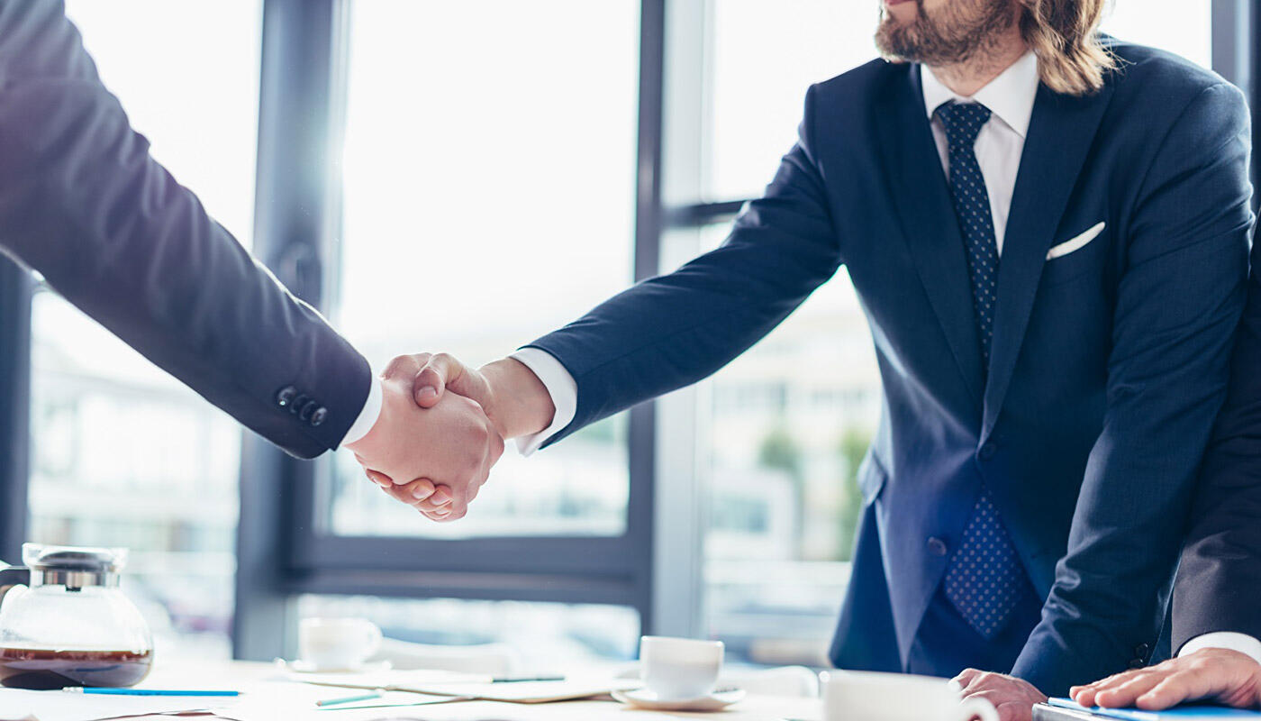 Compliance Zwei Geschäftsleute im Anzug geben sich in einem modernen Büro über einem Konferenztisch die Hand.