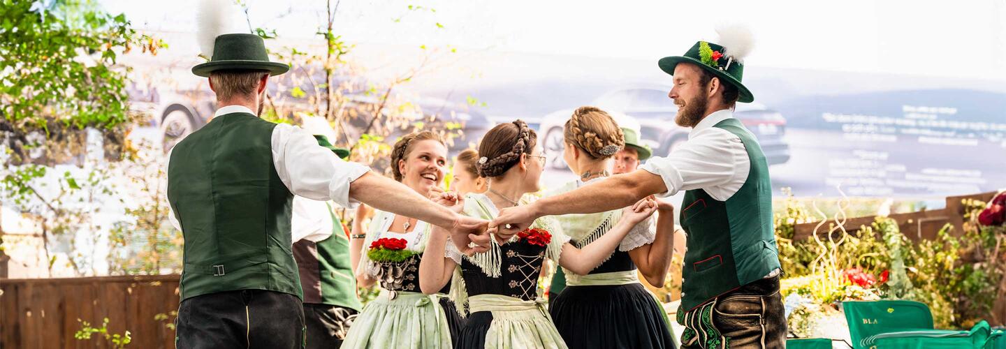 Airbräuwiesn Gruppe von Tänzerinnen und Tänzern in bayerischer Tracht, die sich im Kreis an den Händen halten und fröhlich tanzen. Die Szene wirkt lebhaft und traditionell und spiegelt bayerische Kultur und Volksfeststimmung am Flughafen München wider.