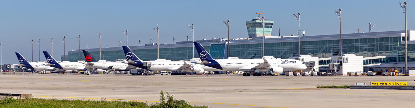 T2 Tochtergesellschaft Mehrere Flugzeuge der Lufthansa stehen am Vorfeld vor dem Terminal 2 des Flughafens München, im Hintergrund ist das Terminalgebäude mit Tower und Abfertigungsbereichen zu sehen.