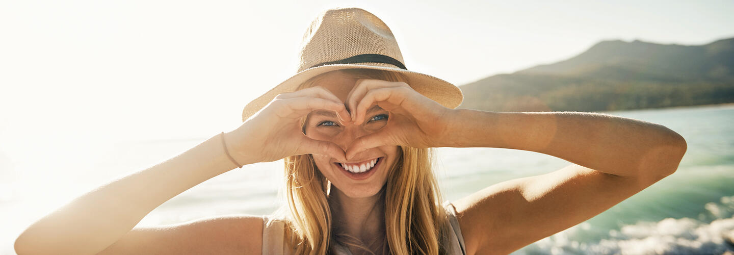 Reiseplanung leicht gemacht Frau am Strand formt mit ihren Händen ein Herz und lächelt in die Kamera, im Hintergrund Meer und Berge.