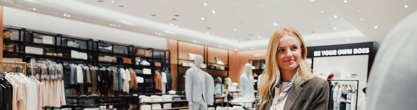 eurotrade Shop Verkäufein Junge Mitarbeiterin in einem modernen Modegeschäft, die lächelnd eine Person berät; im Hintergrund sind Kleiderständer und Schaufensterpuppen zu sehen.