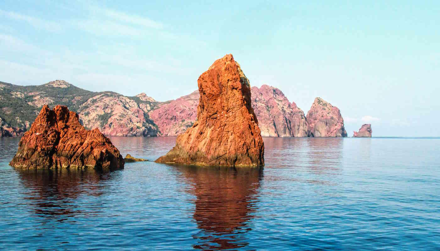 Rote Klippen Korsika – Felsformationen im Mittelmeer Rote Felsen ragen aus dem klaren Wasser vor der Küste Korsikas und spiegeln sich im ruhigen Meer. Im Hintergrund sind weitere zerklüftete Klippen der mediterranen Küstenlandschaft zu sehen.
