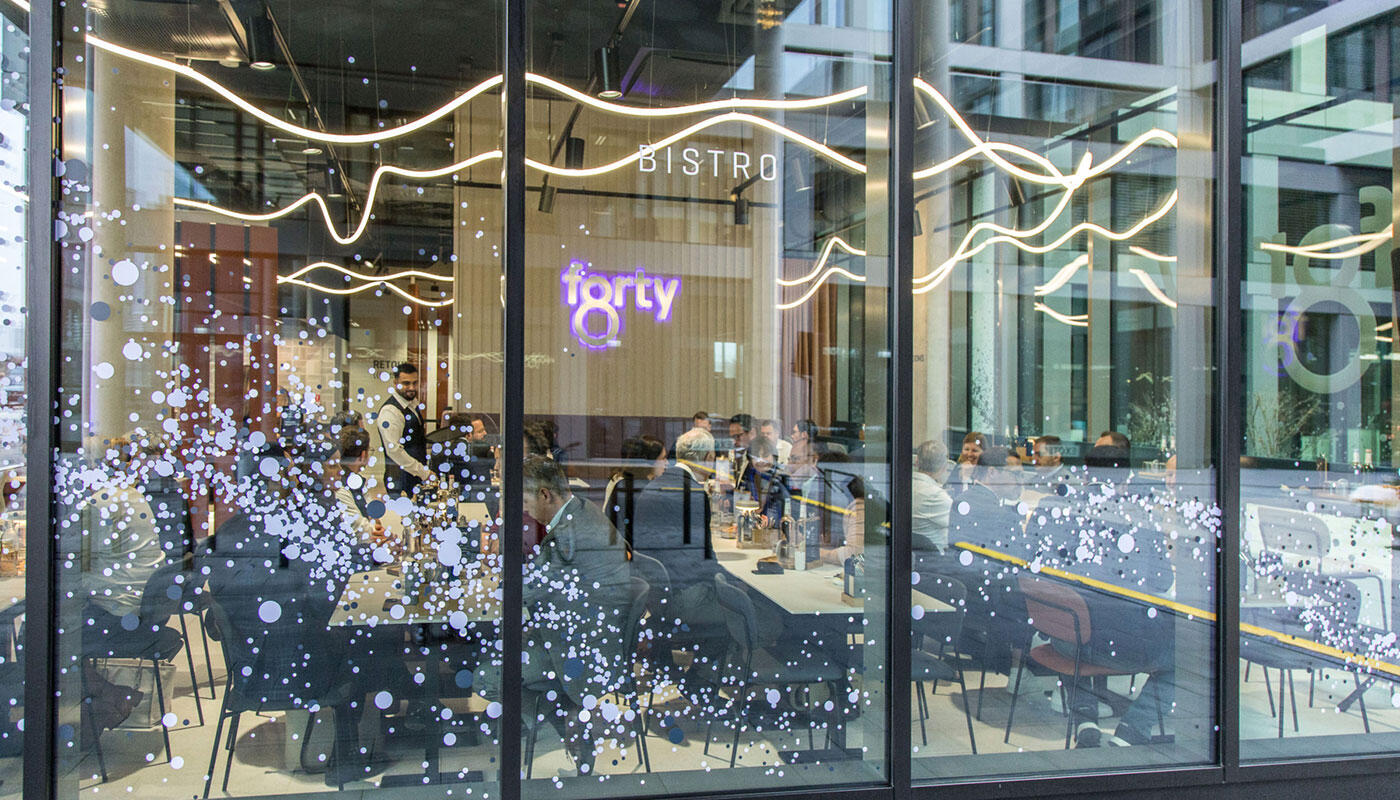 Bistro forty8 Blick von Außen durch die Fensterscheibe des Bistro forty8 im LabCampus. Man sieht ein modern eingerichtetes Bistro mit abendlicher Beleuchtung. Die Stimmung ist entspannt.