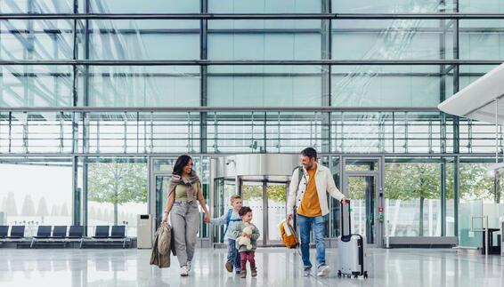 Reisen mit Kindern Mutter und Vater mit zwei kleinen Jungen stehen mit Reisegepäck in einem modernen Flughafengebäude mit Glasfront im Hintergrund