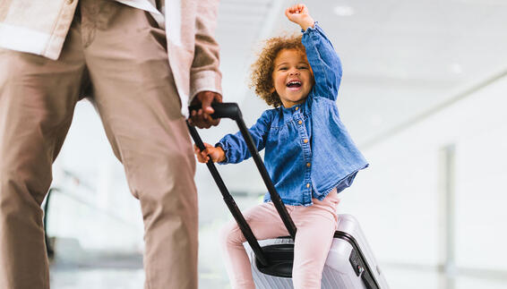 Fliegen mit Kindern Glückliches Kind sitzt auf einem Trolley und wird von Vater gezogen