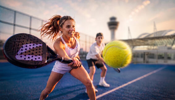 Padel Days am Flughafen München Dynamische Padel-Szene auf Outdoor-Court bei Sonnenuntergang mit Spieler beim kraftvollen Vorhand-Schlag, gelbem Ball im Vordergrund und weiterer Person im Hintergrund – am Flughafen München