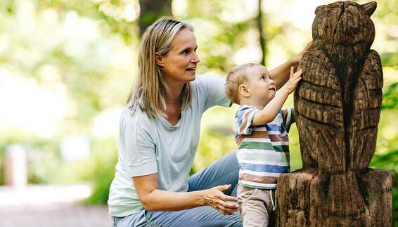Walderlebnispfad im Freisinger Forst Auf dem Walderlebnispfad im Freisinger Forst kniet eine Frau neben einem Kleinkind. Das Kind streckt die Hand aus und berührt eine große geschnitzte Eulenfigur aus Holz.
